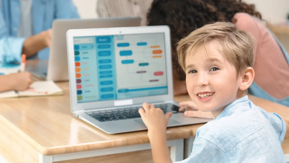 Ein lachender Junge sitzt vor einem Laptop, auf dem ein visuelles Programmierprogramm geöffnet ist. Im Hintergrund arbeiten andere Kinder in einer Gruppenaktivität.