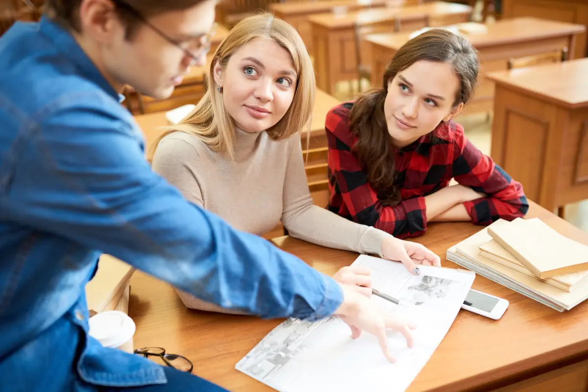 Drei Studierende sitzen an einem Holztisch in einem Klassenzimmer. Eine Person zeigt auf ein Dokument, während die anderen beiden aufmerksam zuhören.