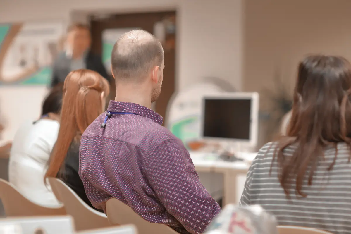 Menschen sitzen in einem Seminarraum und hören einer Präsentation zu. Im Vordergrund ist der Hinterkopf eines Mannes in einem lila Hemd zu sehen, während im Hintergrund ein unscharfes Display und ein Vortragender zu erkennen sind.